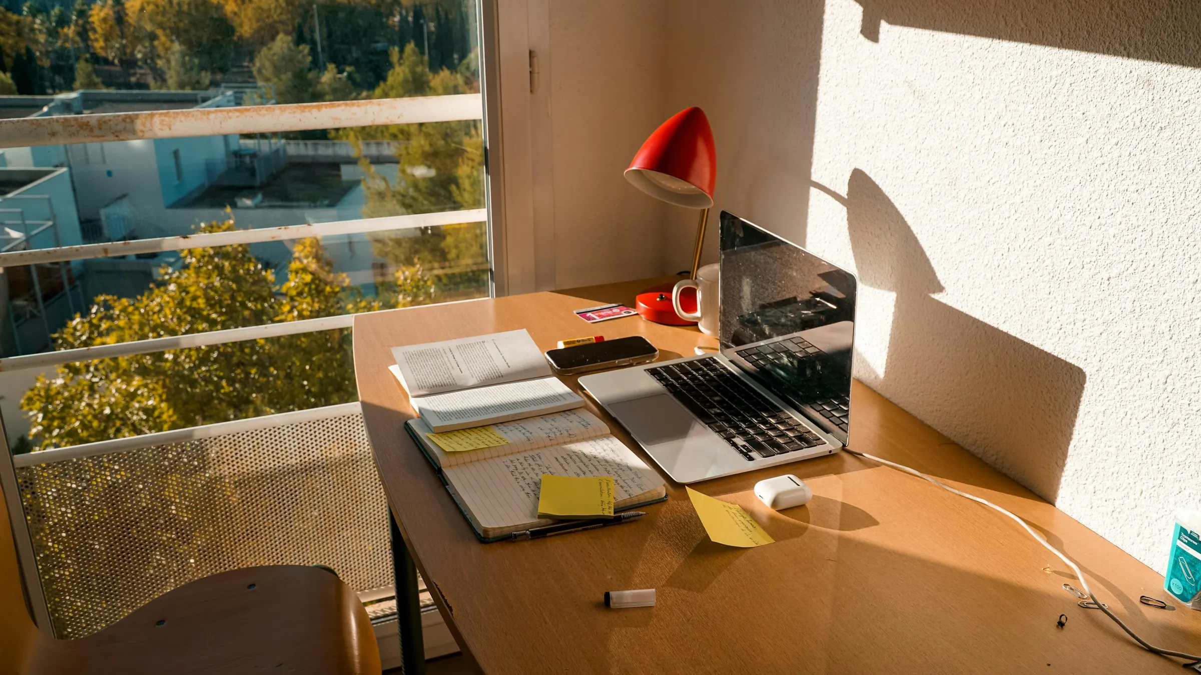A laptop, an open notebook with yellow sticky notes and a red desk lamp on a wooden desk by a sunlit window