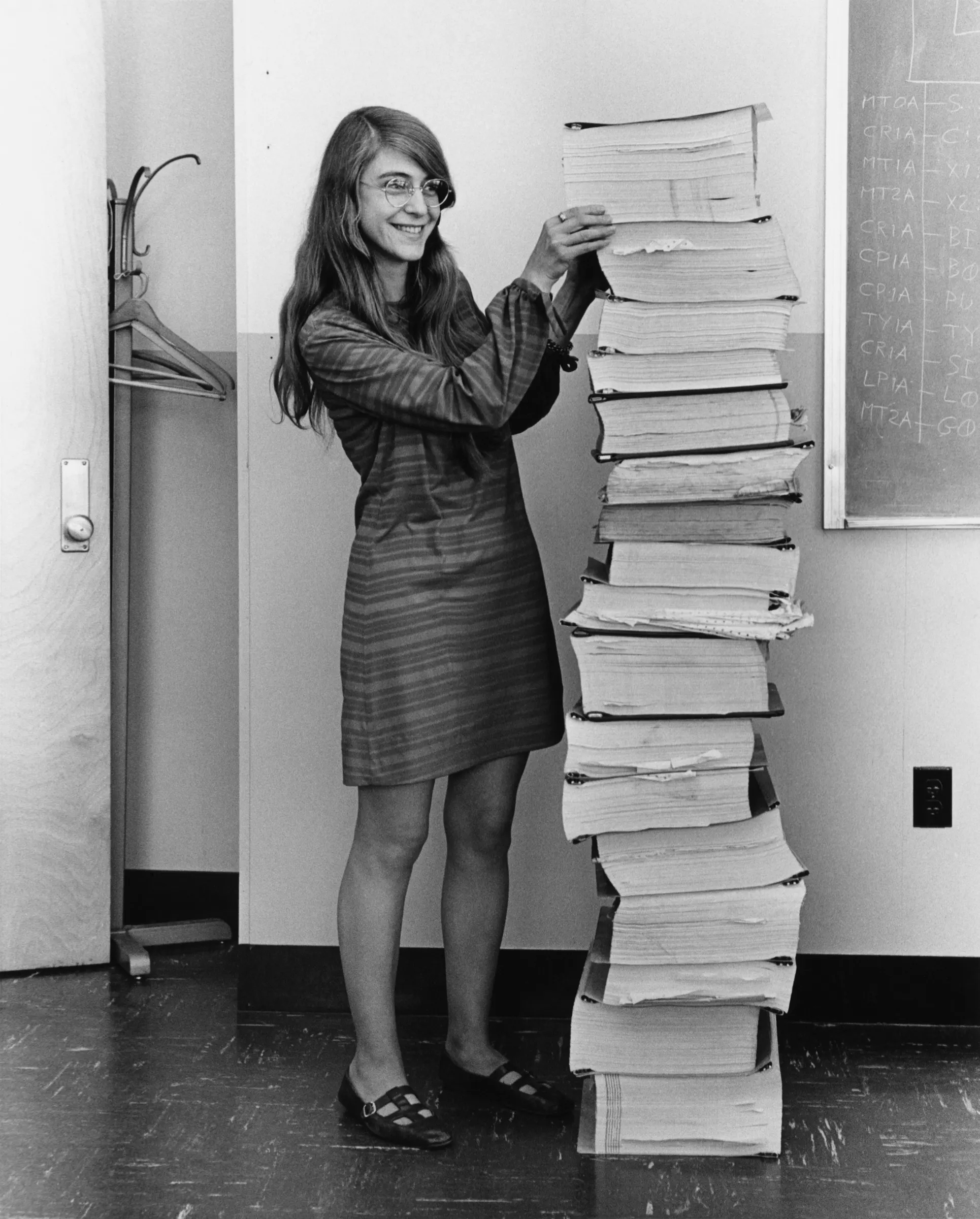 Margaret Hamilton standing beside tall stacks of printed Apollo software listings.